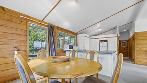 A wooden interior with a round dining table, chairs, and a kitchen area with a microwave and refrigerator. - Seaview, Sun and Surf - Piha Holiday Home (Piha)