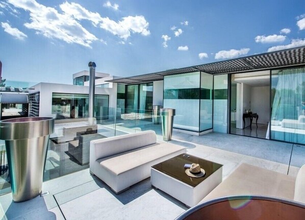 A modern rooftop terrace with glass walls, outdoor seating, and a clear view of the sky.