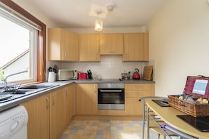 A compact kitchen with wooden cabinets, a built-in oven, a microwave, and a sink. There's a window providing natural light and a table with a basket on it. - Sunnyside Cottage 5 Mins to St Andrews (St. Andrews)