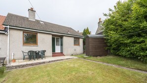 A single-story house with a tiled roof, a front door, and a small front yard with a lawn and a wooden shed.