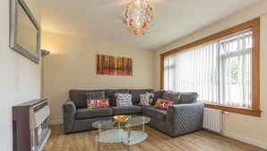 A living room with a sectional sofa, a glass coffee table, a pendant light, a radiator, and a large window with blinds.
