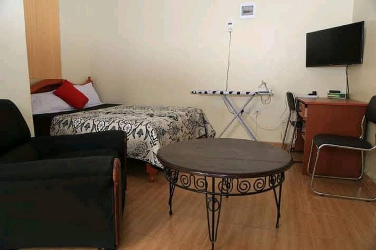 A small, furnished room with a bed, a desk, a chair, and a table.