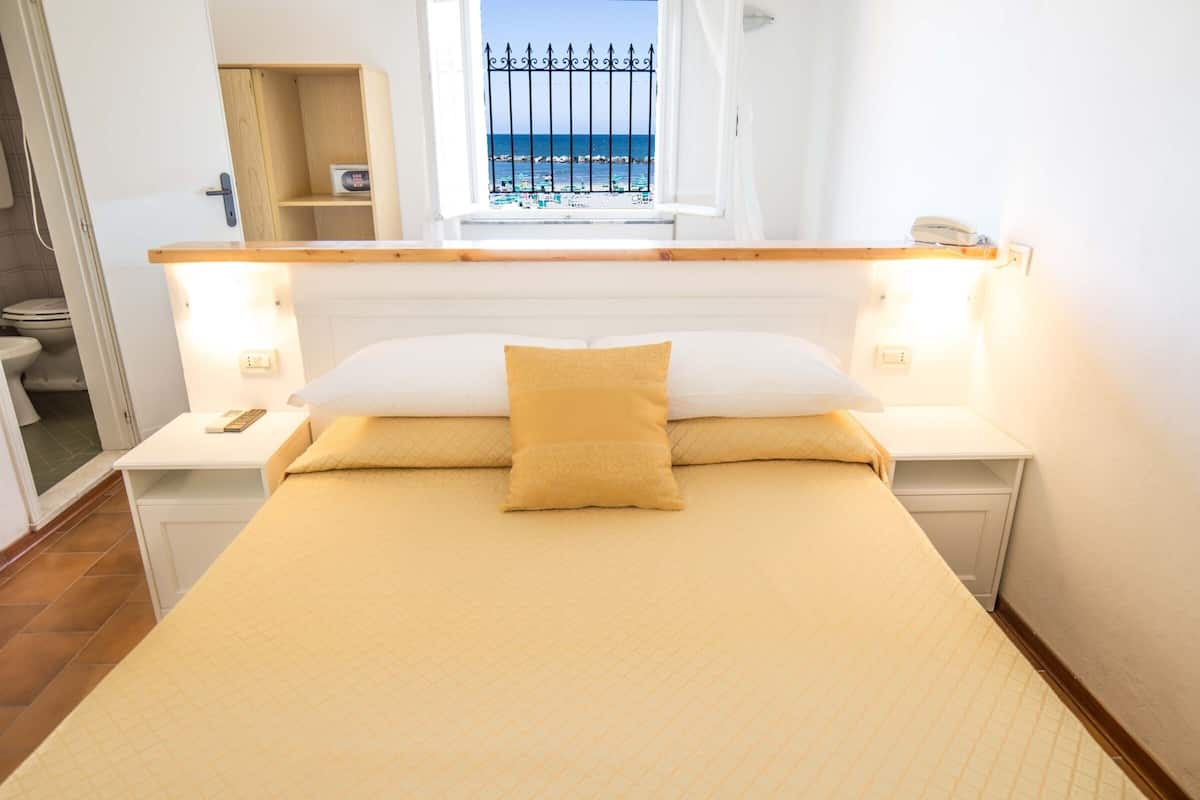 Une chambre à coucher comprenant un lit, des tables de chevet, une commode, une fenêtre donnant sur la mer et une étagère en bois.