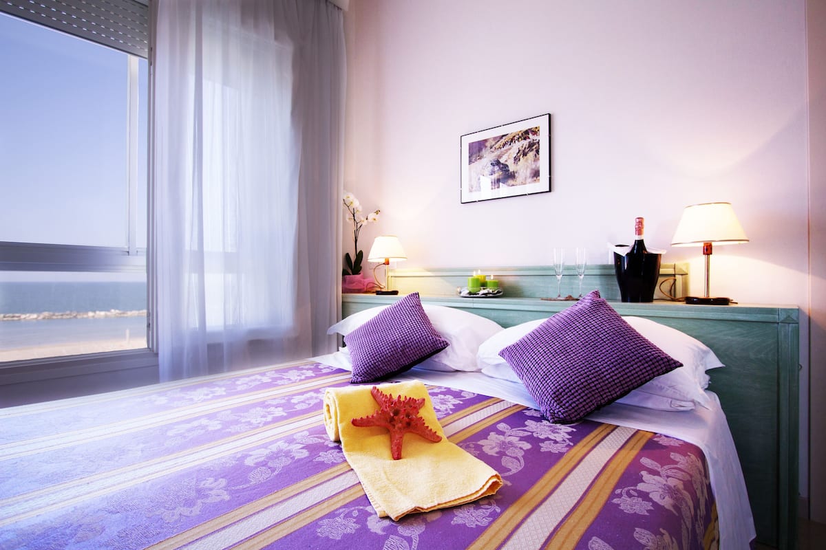 Une chambre d’hôtel avec un lit, deux oreillers, une table de chevet, une lampe, un tableau au mur et une fenêtre donnant sur la mer.