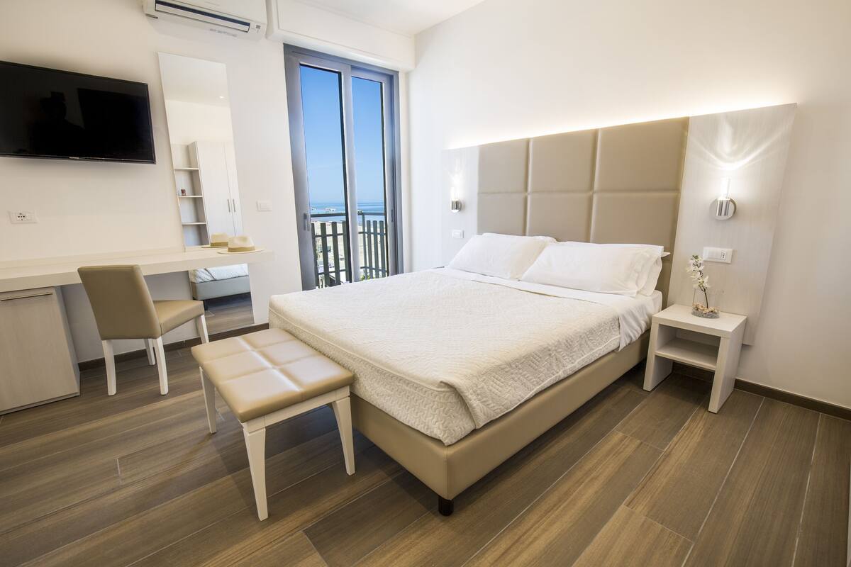 Une chambre d’hôtel moderne dotée d’un grand lit, d’un bureau avec une chaise, d’un téléviseur fixé au mur et d’un balcon offrant une vue sur la mer.