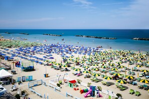 Ein Strand mit vielen Sonnenschirmen und Liegestühlen, einem Empfangsgebäude und einem Spielplatz.