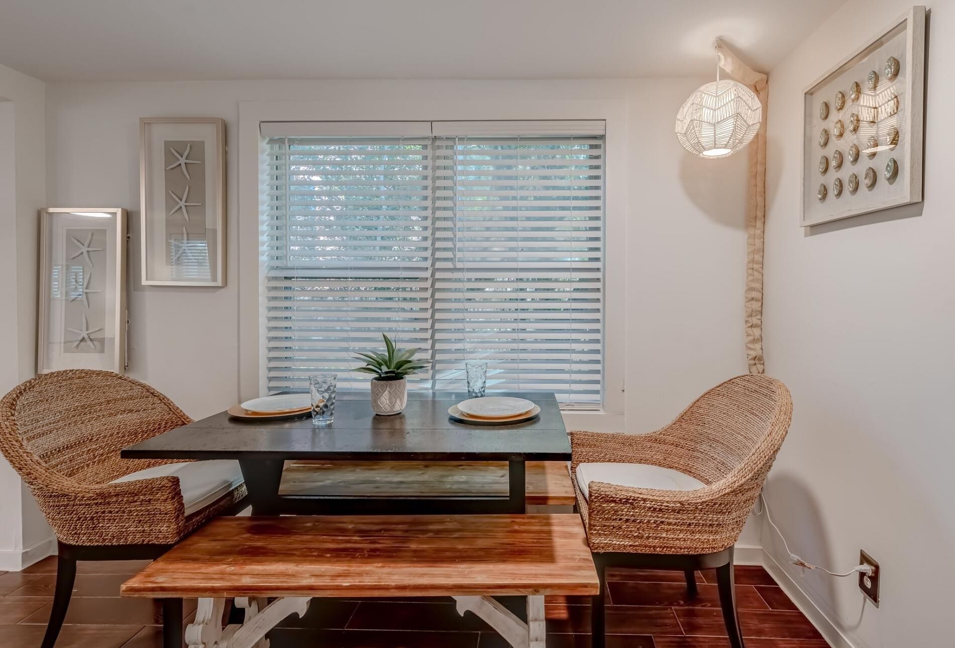 A dining area with a wooden table and woven chairs, a large window with blinds, and a pendant light.