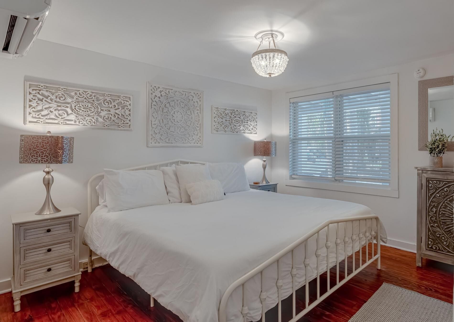 A bedroom with a white bed, decorative wall panels, a chandelier, and a window with blinds.