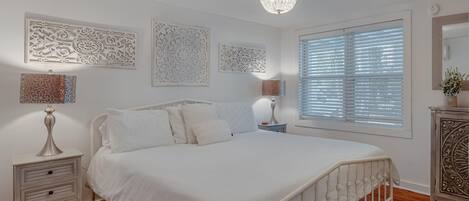 A bedroom with a white bed, decorative wall panels, a chandelier, and a window with blinds.