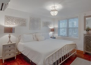 A bedroom with a white bed, decorative wall panels, a chandelier, and a window with blinds. - Charming 1950s Beach Cottage Apt B (Isle of Palms)