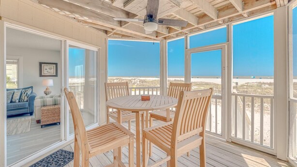 Un espacio exterior techado con una mesa y sillas de madera, un ventilador de techo y vista a la playa.
