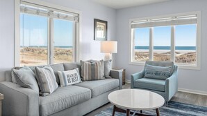 Un salón con un sofá gris, una mesita de centro redonda y un sillón azul. Hay ventanas grandes con vistas a la playa y al océano.