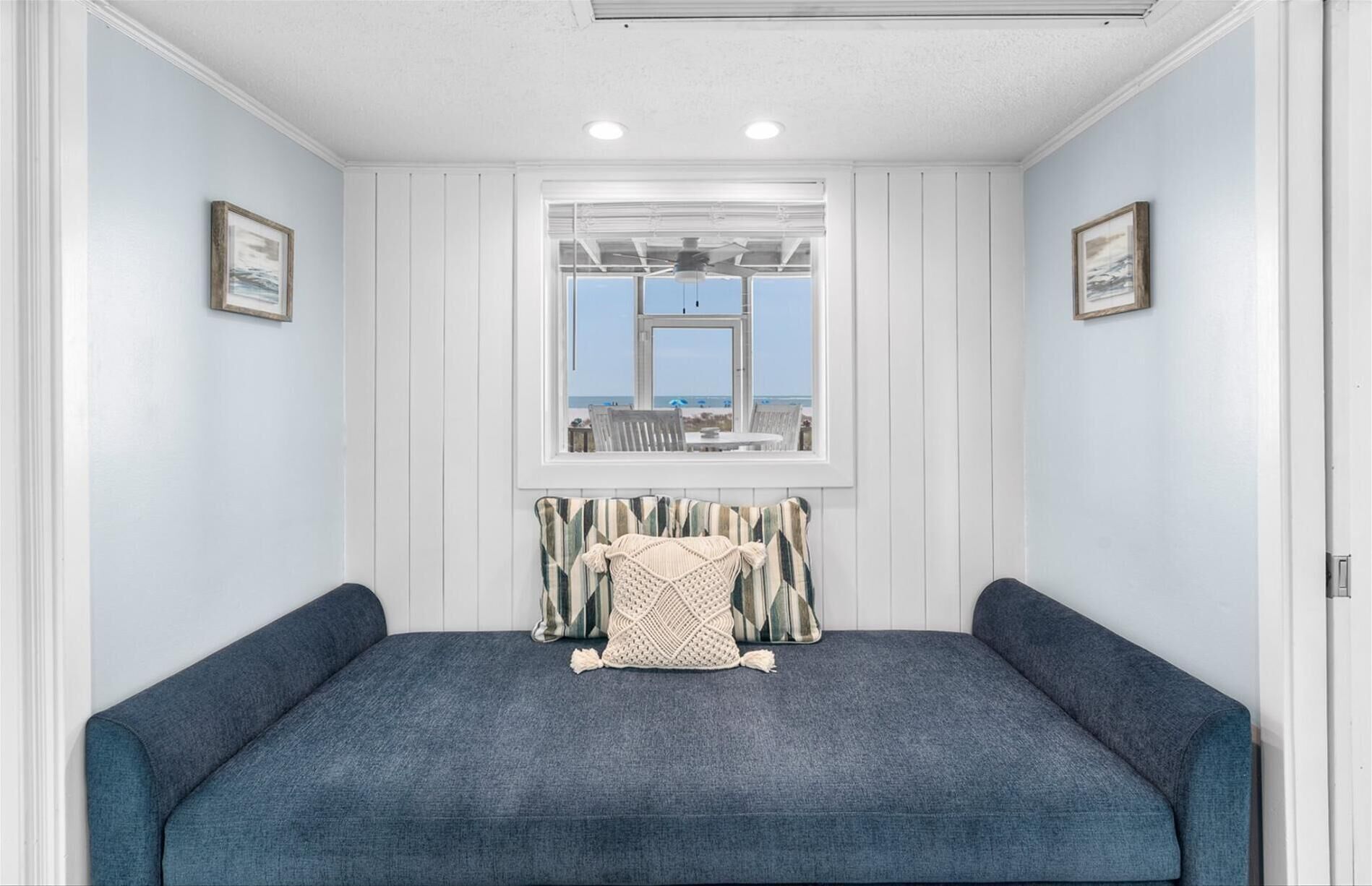 A room with a large window offering a view of the beach, a blue sofa, and a patterned throw pillow.