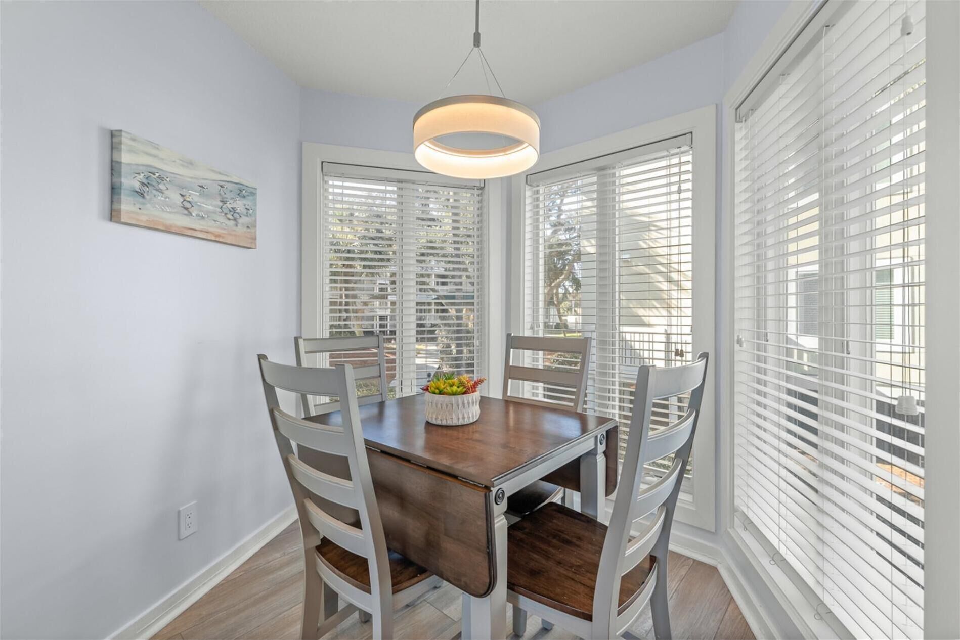 Un comedor con una mesa de madera y cuatro sillas, una lámpara colgante y persianas blancas en las ventanas.