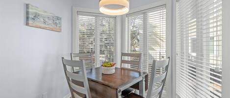 Un comedor con una mesa de madera y cuatro sillas, una lámpara colgante y persianas blancas en las ventanas.