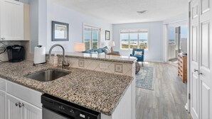 Una cocina moderna con encimeras de granito, lavavajillas y cafetera. Hay un comedor con una silla azul y vistas a la playa a través de grandes ventanales.