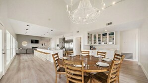 A spacious dining area with a round wooden table and chairs, a modern chandelier, and a kitchen in the background. - Beachfront Beauty with Huge Deck & Stunning Views (Galveston)