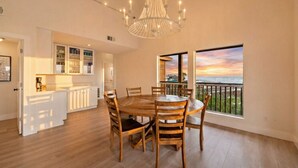 A spacious dining area with a large wooden table and chairs, a chandelier, and a view of the sunset through large windows. - Beachfront Beauty with Huge Deck & Stunning Views (Galveston)