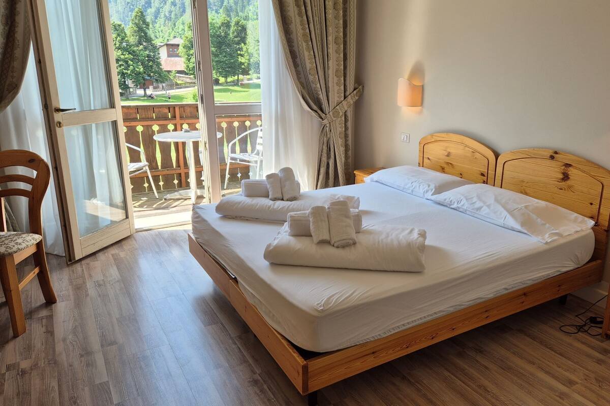 A double bed with white linens and towels, wooden headboard, bedside table, and a view of a wooden deck and greenery outside.