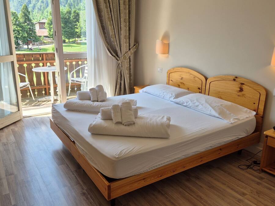 Cama doble con ropa de cama y toallas blancas, cabecera de madera, mesita de noche y vistas a una terraza de madera y follaje exterior.