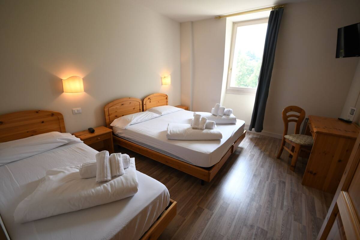 A hotel room with two beds, wooden headboards, bedside tables, a desk, a chair, and a window with curtains.