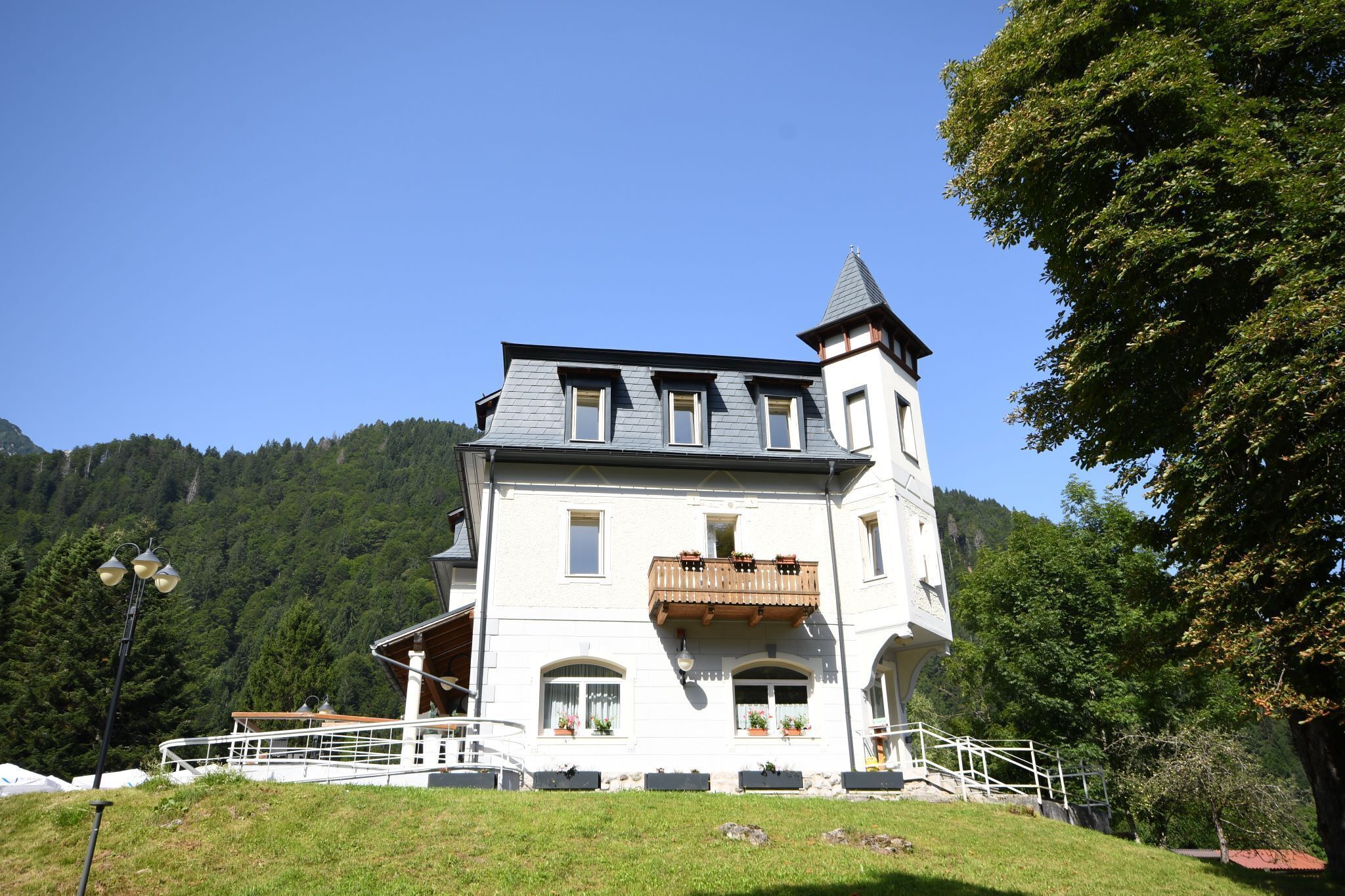 A large, white building with a tower and a balcony, surrounded by greenery and mountains.