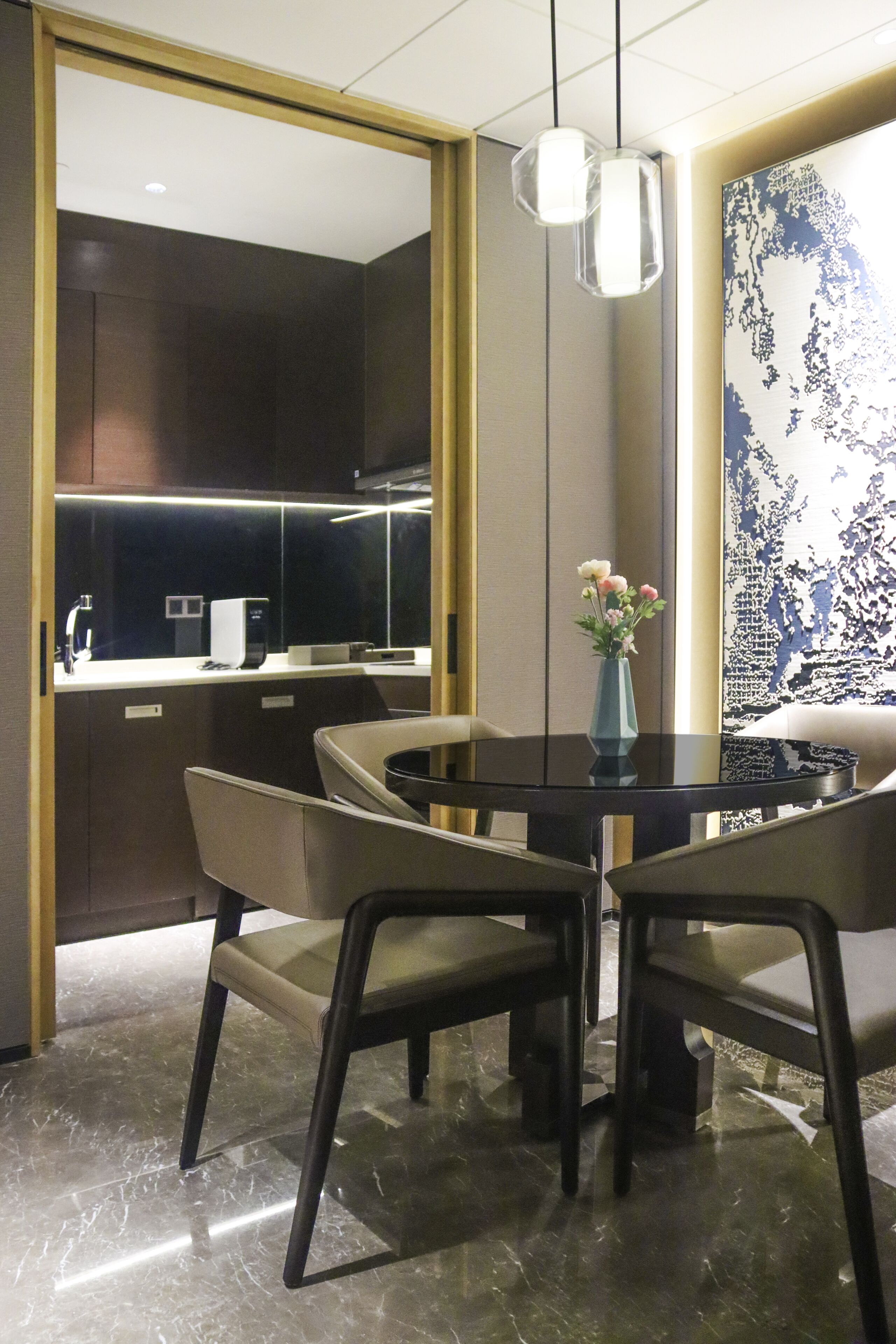 A modern dining area with a glass table, four chairs, a vase with flowers, and a view of a kitchen and living area through a glass partition.