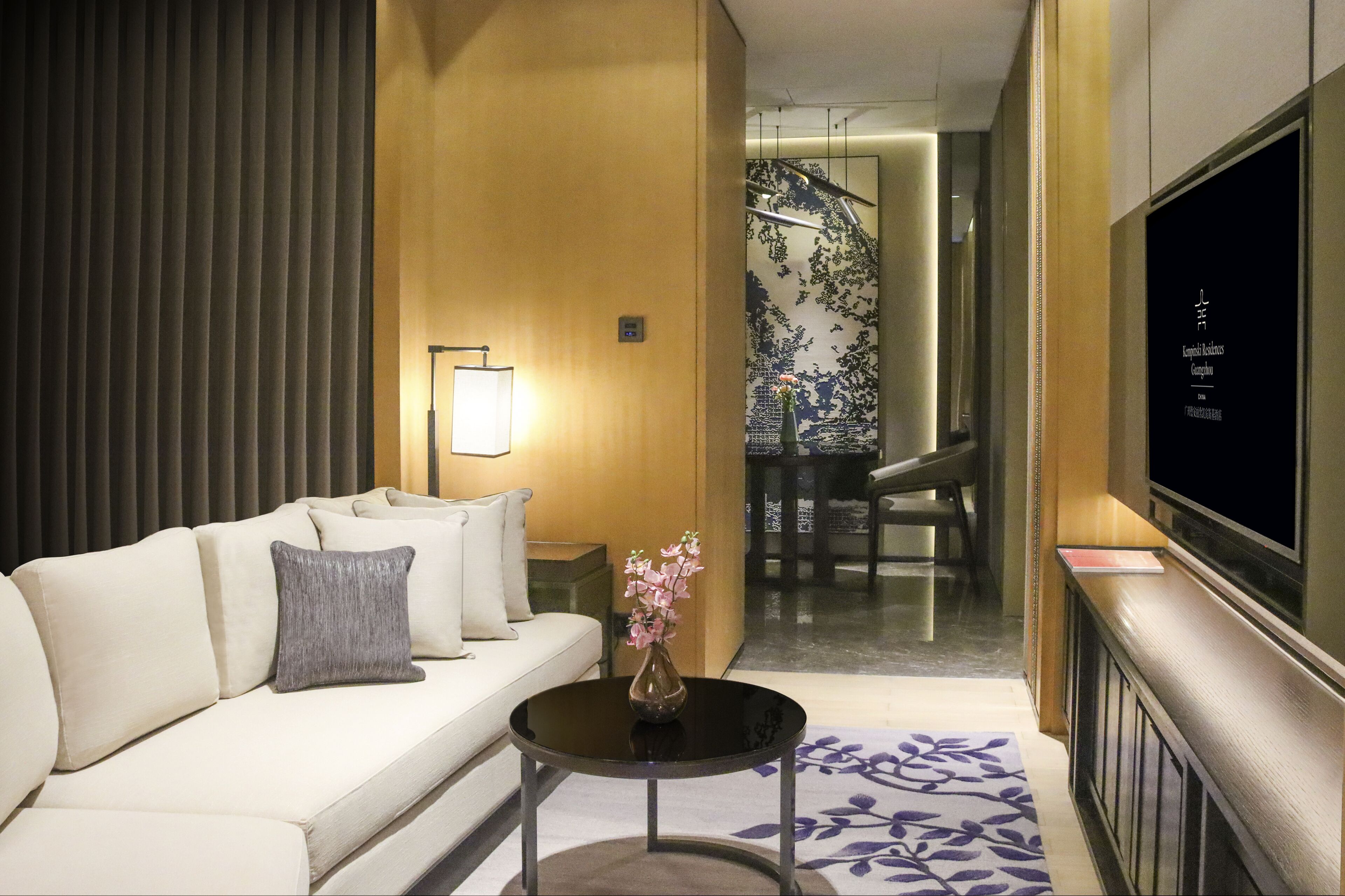 A modern hotel room with a sofa, a small round table with a vase, a flat-screen TV, and a patterned rug.