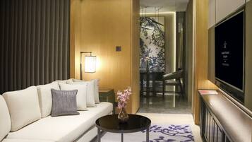 A modern hotel room with a sofa, a small round table with a vase, a flat-screen TV, and a patterned rug.