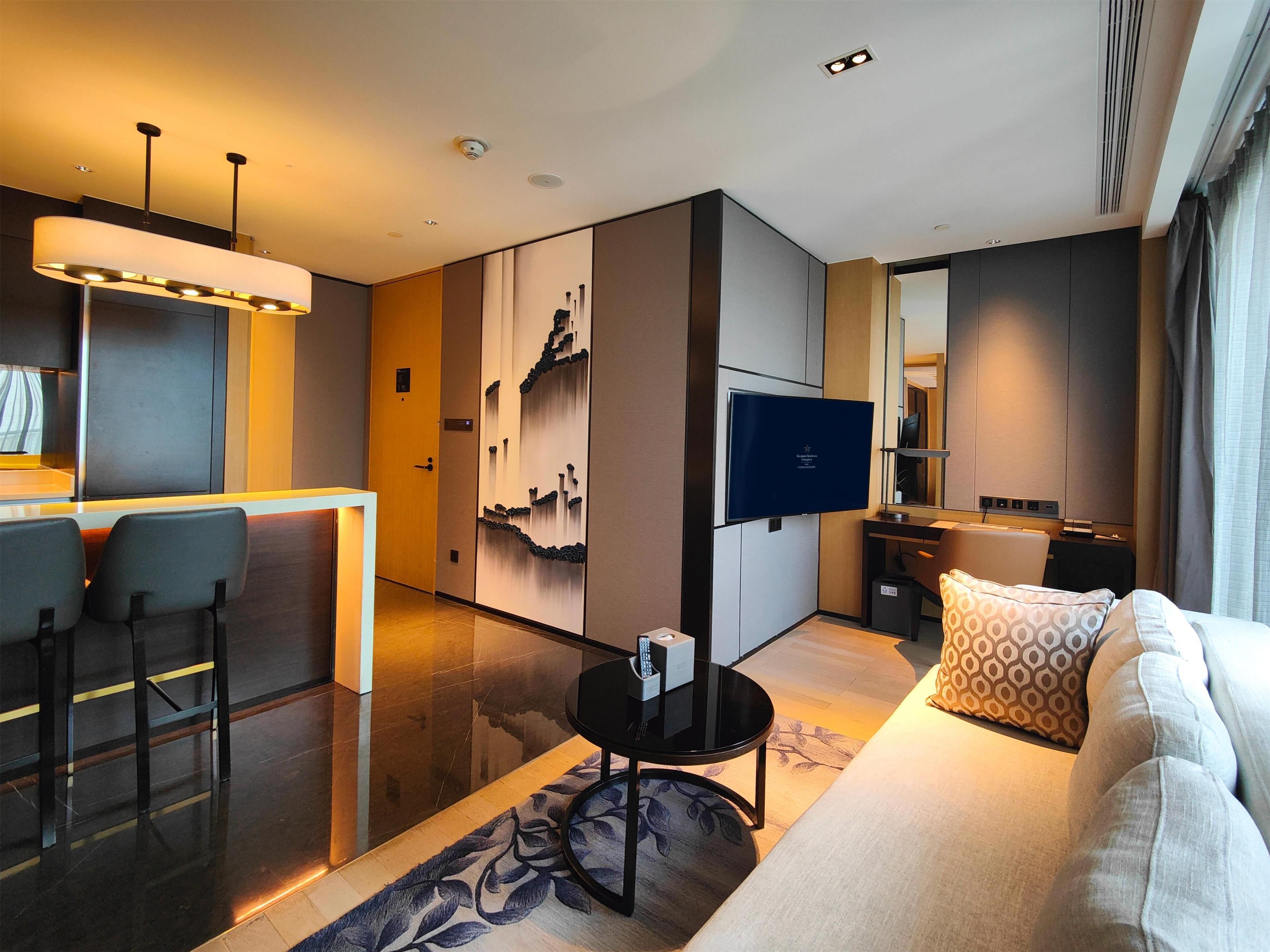 A modern hotel room with a sofa, a TV, a small table, and a desk area.
