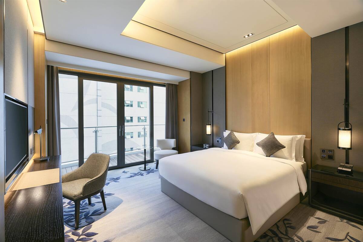 Quarto de hotel moderno com uma cama grande, uma escrivaninha, uma cadeira e vista da cidade por amplas janelas.