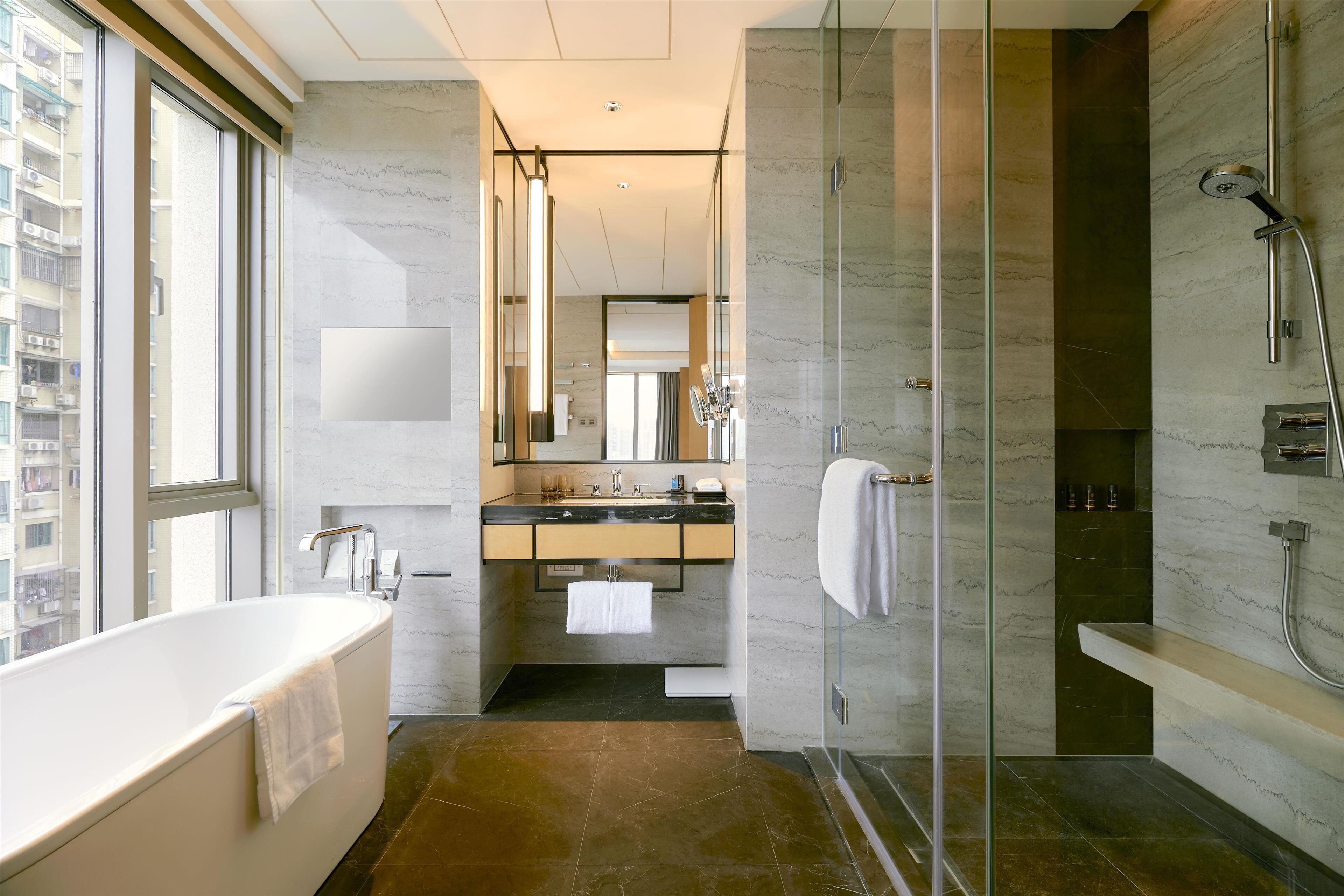 Ein modernes Badezimmer mit Badewanne, einer Duschkabine, einem Waschtisch mit Spiegel und Blick auf die Stadt durch große Fenster.