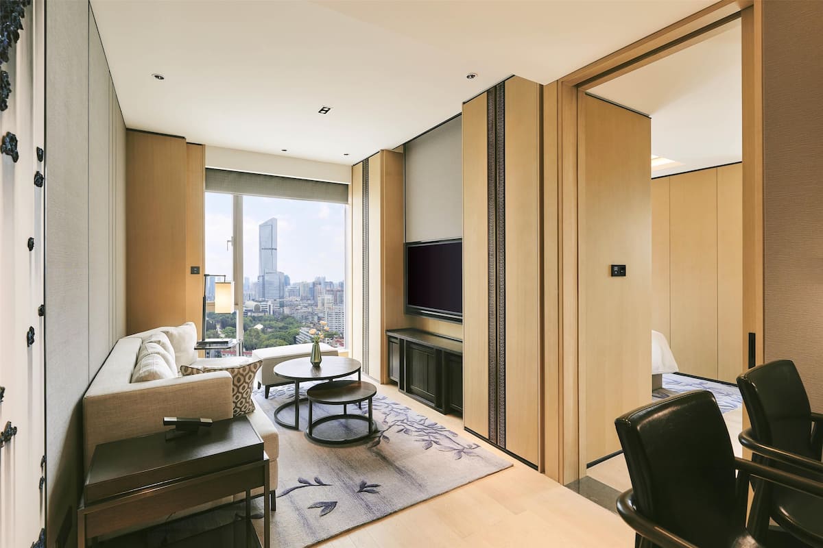 Quarto de hotel moderno com sofá, uma mesa redonda pequena, TV de tela plana e vista para a paisagem urbana.