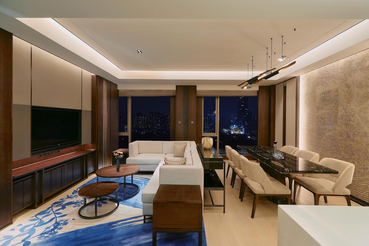Uma sala de estar moderna com uma televisão de tela plana, um sofá, uma mesa de jantar com cadeiras e uma janela grande com vista para a cidade à noite.