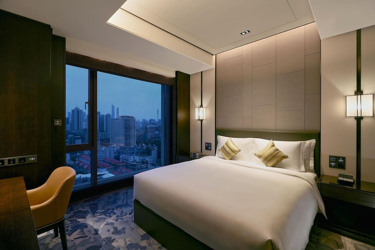 Quarto de hotel com uma cama grande, uma escrivaninha e uma cadeira. Vê-se uma vista da cidade através de amplas janelas.