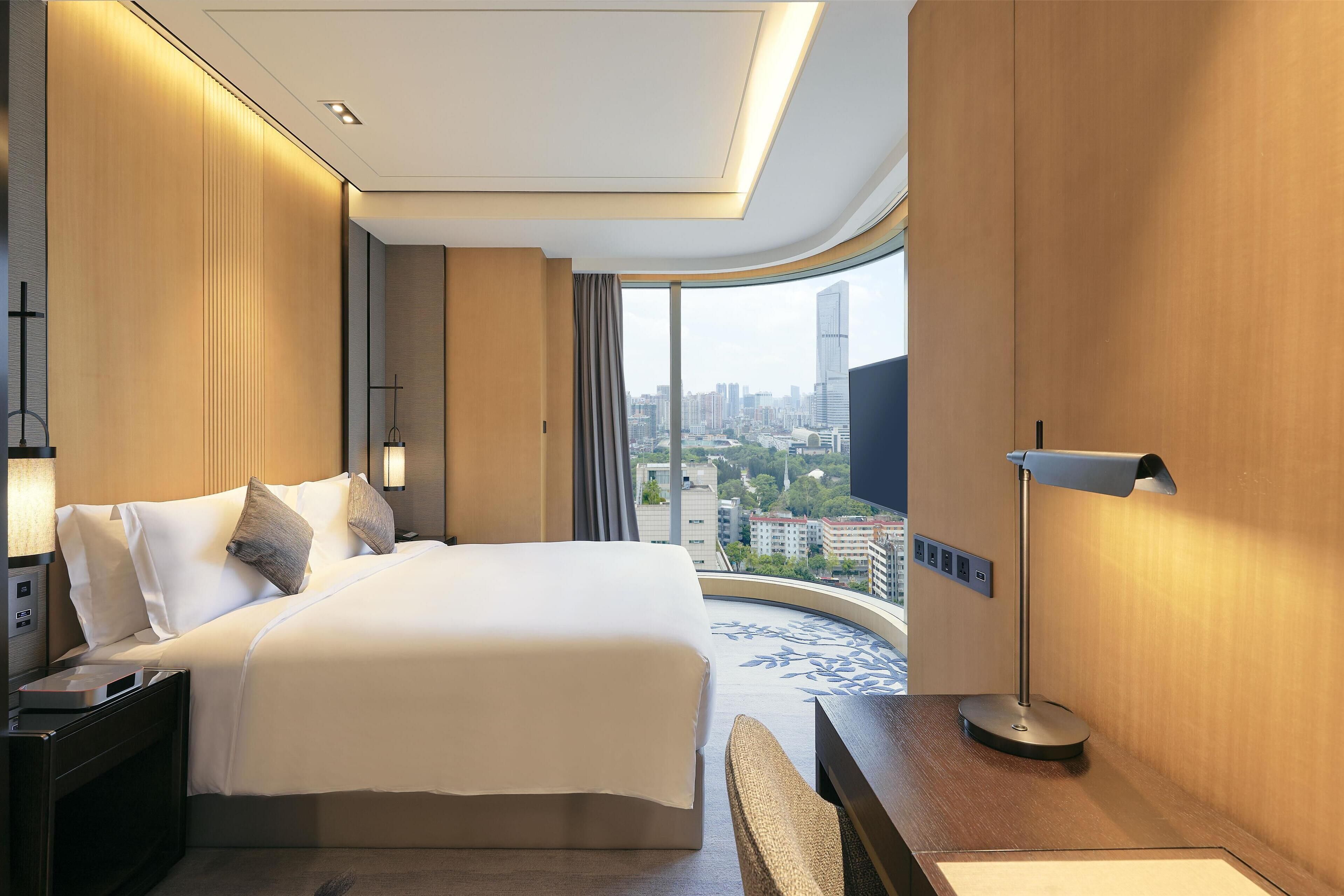 A modern hotel room with a large bed, bedside tables, a desk, and a view of the city through a large window.