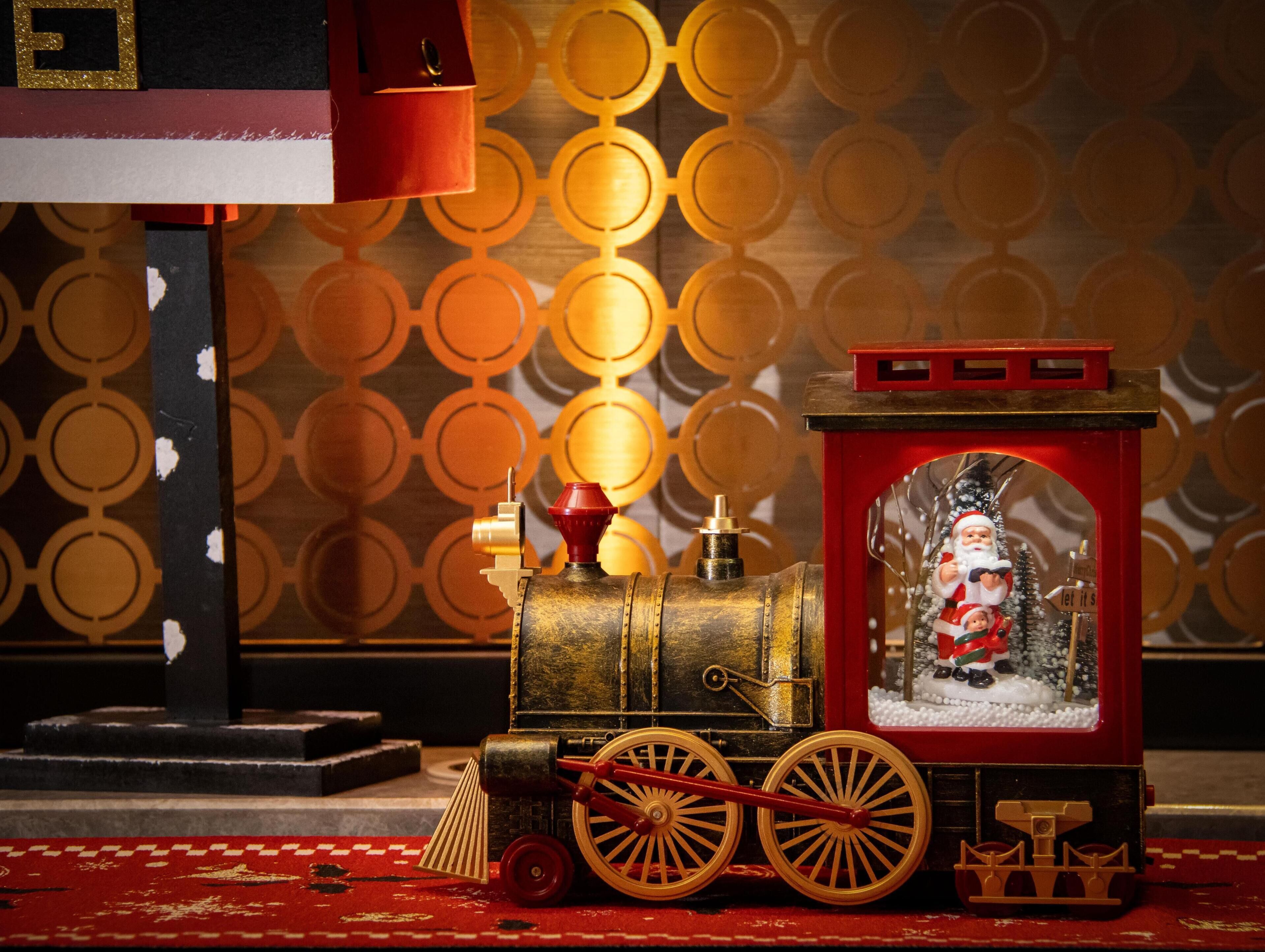 Un tren modelo con una figura de Papá Noel en la ventana, frente a un papel tapiz estampado.