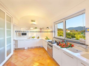 Uma cozinha moderna com armários brancos, forno embutido e janela com vista para vegetação e colinas.