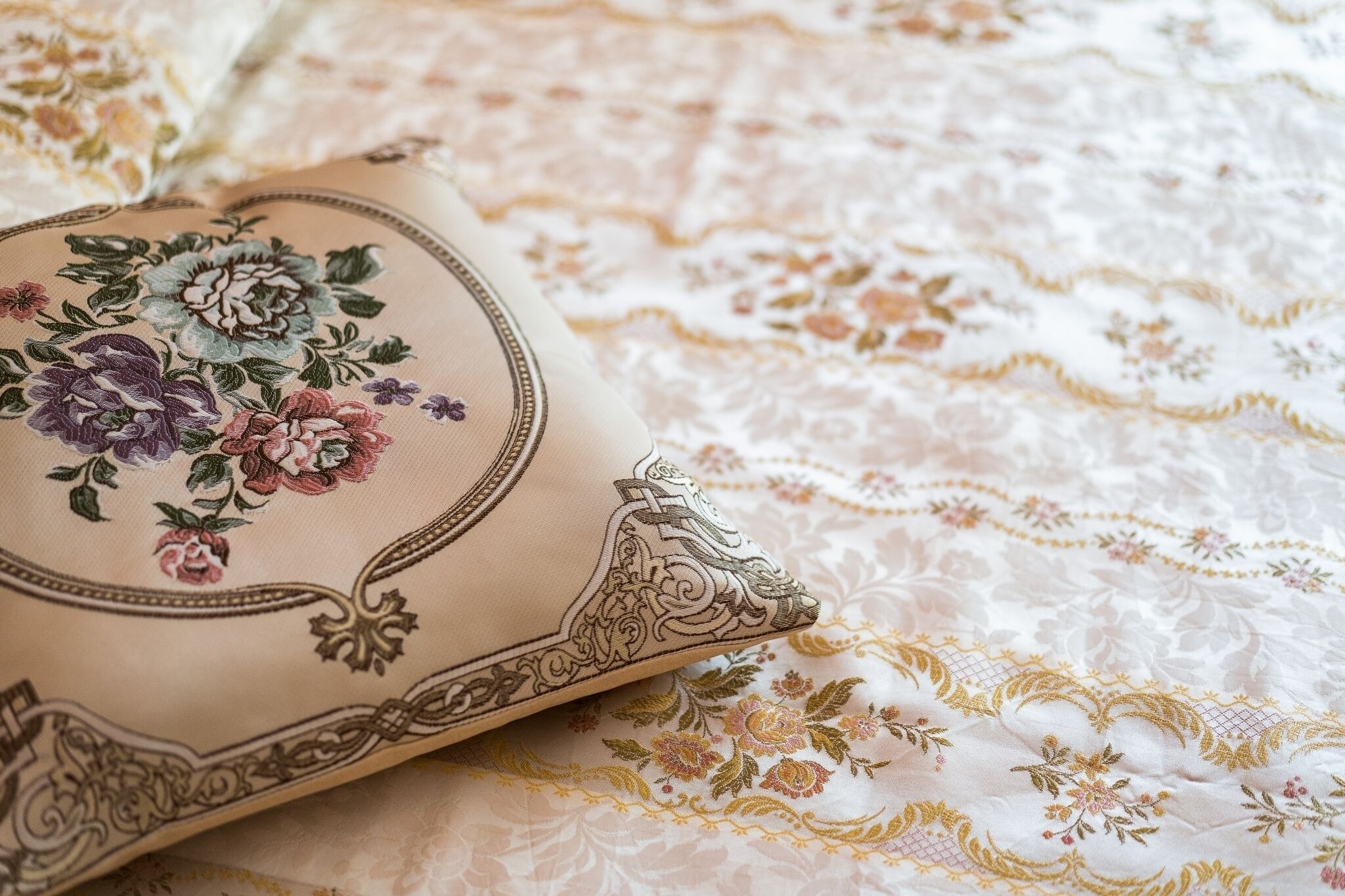 A beige pillow with floral embroidery on a bed with a white and gold floral patterned bedspread.