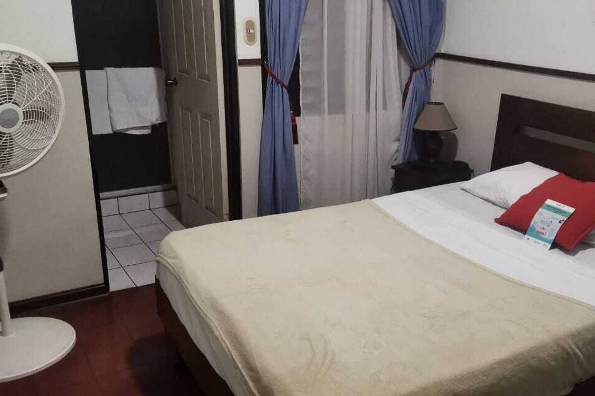 Une chambre d’hôtel avec un lit, une table de chevet avec une lampe, un ventilateur sur pied et une porte avec des rideaux.