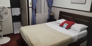 A hotel room with a bed, a bedside table with a lamp, a standing fan, and a door with curtains. - Hotel Santamaria (Alajuela)