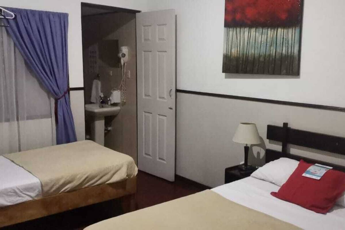 Une chambre d’hôtel avec deux lits, une table de chevet avec une lampe, un tableau au mur et une salle de bain visible à travers une porte ouverte.