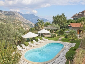 A swimming pool with lounge chairs and umbrellas, surrounded by lush greenery and mountains in the background.