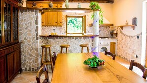 Une cuisine rustique aux murs de pierre, avec des tabourets de bar en bois et une table à manger en bois sur laquelle se trouve un panier de fruits.