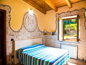 A bedroom with a stone wall, a bed with a striped bedspread, a stone fireplace, and a window with a view of greenery.