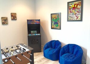Quarto com mesa de futebol de mesa, dois pufes azuis e posters de histórias em quadrinhos na parede.
