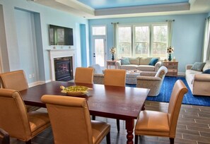 A dining area with a wooden table and chairs, a fireplace, a television mounted above it, and a living room with a sofa and a coffee table.