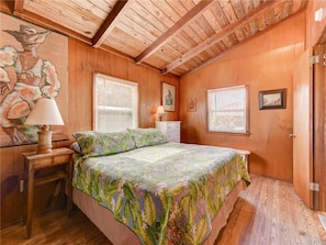 Un dormitorio con paredes de madera, una cama con colcha estampada, una mesita de noche con lámpara y un cuadro en la pared.