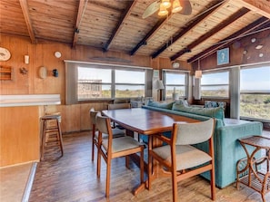 A wooden interior with a dining table, chairs, and a sofa. There are large windows providing a view of the outdoors. - Pounds Cottage, 1 Bedroom, Ocean Front, Pet Friendly, Sleeps 5 (St. Augustine)