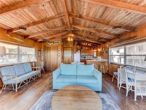 Interior rústico de una cabaña con techo de madera, sofá azul claro, mobiliario de mimbre y una cocina visible a través de una puerta abierta.
